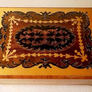 Puede incluir: Una caja de música de madera con un diseño de incrustación decorativo que presenta patrones florales negros y dorados sobre un fondo marrón claro.
