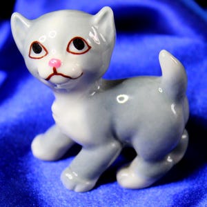 May include: A small, gray and white ceramic figurine of a kitten. The kitten has brown eyes, a pink nose, and a sweet expression.