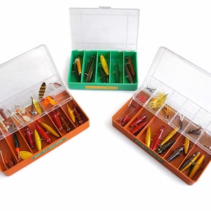 May include: Three open tackle boxes filled with fishing lures. Two orange boxes and one green box are visible, each with clear lids. The lures are various colors, including red, yellow, and silver, and are arranged in separate compartments.