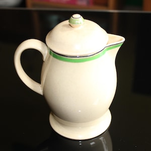 May include: Cream-colored ceramic creamer with a lid, handle, and spout. The creamer has a green and black band around the top and a small green knob on the lid. The creamer sits on a small, round base.