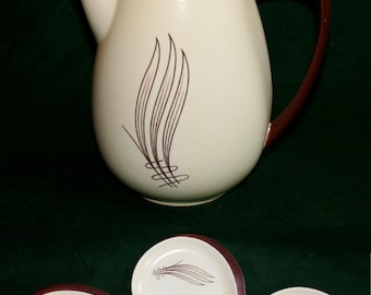 Może przedstawiać: Kremowy ceramiczny dzbanek do kawy z brązową pokrywką i uchwytem. Dzbanek ma stylizowany wzór liścia w kolorze brązowym. Trzy pasujące kremowe ceramiczne spodki z brązowymi obwódkami i stylizowanym wzorem liścia w kolorze brązowym.