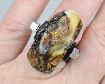 May include: A ring with a large, amber-colored stone set in a black metal band. The stone is irregularly shaped and has a swirling pattern of brown and yellow.