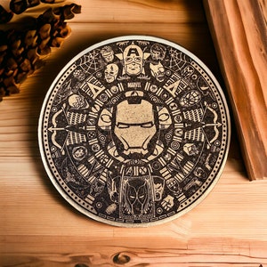 Puede incluir: Un posavasos redondo de madera con un diseño en blanco y negro que presenta personajes de Marvel. El centro del posavasos presenta la máscara de Iron Man. El posavasos tiene la palabra "MARVEL" impresa.
