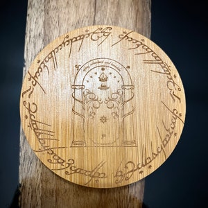 Pode incluir: Um coaster redondo de madeira com uma imagem gravada da entrada das Minas de Moria do Senhor dos An&eacute;is. A inscri&ccedil;&atilde;o ao redor da borda diz: "Um Anel para govern&aacute;-los a todos, Um Anel para encontr&aacute;-los, Um Anel para traz&ecirc;-los a todos e nas trevas prend&ecirc;-los."
