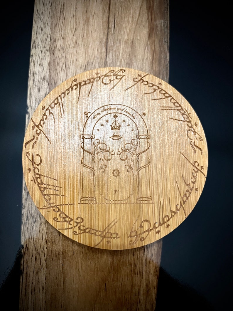 Pode incluir: Um coaster redondo de madeira com uma imagem gravada da entrada das Minas de Moria do Senhor dos An&eacute;is. A inscri&ccedil;&atilde;o ao redor da borda diz: "Um Anel para govern&aacute;-los a todos, Um Anel para encontr&aacute;-los, Um Anel para traz&ecirc;-los a todos e nas trevas prend&ecirc;-los."