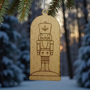 May include: Wooden Christmas ornament featuring a laser-etched nutcracker design. The ornament is rectangular with a rounded top and a small hole for hanging. The nutcracker is depicted in a traditional uniform with a hat and mustache. The background is a snowy forest.
