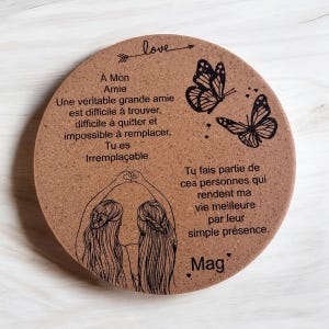 May include: Cork coaster featuring a black and white illustration of two women holding hands and forming a heart shape with their fingers. The text "A Mon Amie" and "love" are engraved on the coaster, along with a message about friendship. Two black butterflies are also engraved on the coaster.