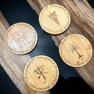 Pode incluir: Quatro  apoios de copo redondos de madeira com um design em preto e branco que apresenta a inscri&ccedil;&atilde;o "Um Anel para govern&aacute;-los a todos, Um Anel para encontr&aacute;-los, Um Anel para traz&ecirc;-los a todos e nas trevas prend&ecirc;-los" e v&aacute;rios s&iacute;mbolos de O Senhor dos An&eacute;is.