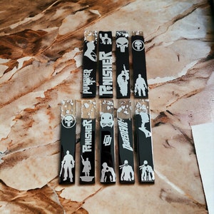May include: A collection of black resin bookmarks featuring white superhero-themed designs. The bookmarks display characters and logos, including "Punisher" and "Daredevil". Some bookmarks have silver leaf accents.