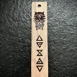 May include: Wooden bookmark with a wolf head and a symbol of four triangles. The bookmark is brown and has a hole at the top.