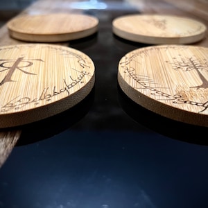 Pode incluir: Quatro  t&aacute;buas redondas de madeira com acabamento marrom escuro. As t&aacute;buas apresentam desenhos gravados a laser, incluindo a inscri&ccedil;&atilde;o "Um Anel para govern&aacute;-los a todos" e uma &aacute;rvore com estrelas.
