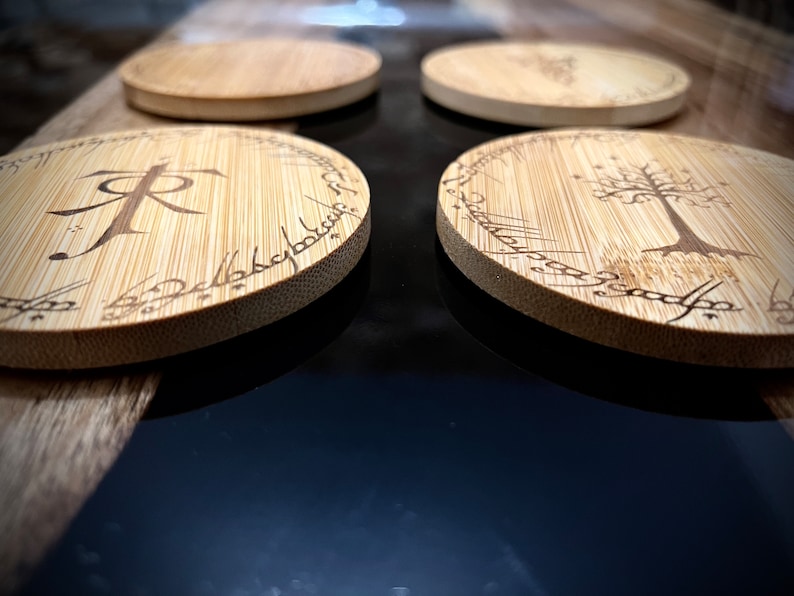 Pode incluir: Quatro  t&aacute;buas redondas de madeira com acabamento marrom escuro. As t&aacute;buas apresentam desenhos gravados a laser, incluindo a inscri&ccedil;&atilde;o "Um Anel para govern&aacute;-los a todos" e uma &aacute;rvore com estrelas.
