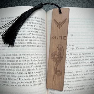 Puede incluir: Un marcapáginas de madera con un diseño marrón en forma de serpiente y la palabra "DUNE" en letras blancas. El marcapáginas está decorado con un símbolo estilizado de un gusano de arena.