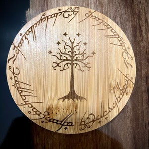 Pode incluir: Um descanso de copo redondo de madeira com um design gravado a laser da &Aacute;rvore Branca de Gondor de O Senhor dos An&eacute;is. A inscri&ccedil;&atilde;o em torno da &aacute;rvore diz "Um Anel para govern&aacute;-los a todos, Um Anel para encontr&aacute;-los, Um Anel para traz&ecirc;-los a todos e nas trevas prend&ecirc;-los."
