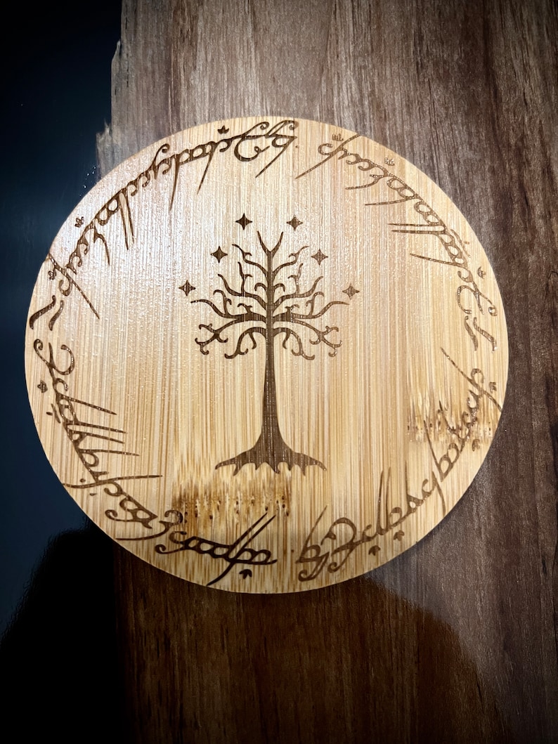 Pode incluir: Um descanso de copo redondo de madeira com um design gravado a laser da &Aacute;rvore Branca de Gondor de O Senhor dos An&eacute;is. A inscri&ccedil;&atilde;o em torno da &aacute;rvore diz "Um Anel para govern&aacute;-los a todos, Um Anel para encontr&aacute;-los, Um Anel para traz&ecirc;-los a todos e nas trevas prend&ecirc;-los."
