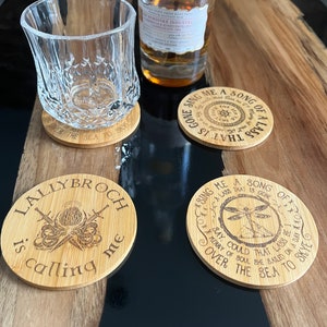 May include: Four round wooden coasters with different designs. The coasters feature a dragonfly, a compass, a thistle, and a quote about a song of a lass. The coasters are made of light brown wood and have a natural finish.
