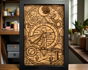 May include: A framed wooden art piece with a black frame. The artwork features a detailed laser-cut design with a central figure, text, a rose, a snake, and a crescent moon. The color is a light brown.