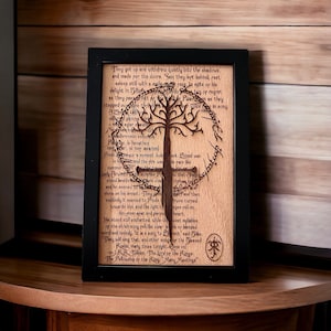 Puede incluir: Obra de arte enmarcada en madera con un diseño de árbol cortado con láser y texto de "El Señor de los Anillos". La obra de arte está en un marco negro y descansa sobre una superficie de madera. El texto incluye el nombre del autor y el título del libro.