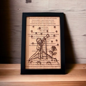 May include: A wooden plaque with a black frame features a laser-cut design of a small prince looking through a telescope at a star-filled sky. The text on the plaque reads: "Le petit prince arracha..." and continues with a passage in French. The plaque is a decorative piece for a home or office.