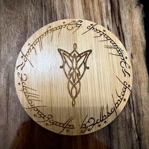 Pode incluir: Um descanso de copo redondo de madeira com acabamento em gr&atilde;o de madeira castanho. O descanso de copo apresenta um design gravado a laser do Um Anel de O Senhor dos An&eacute;is, com a inscri&ccedil;&atilde;o "Um Anel para govern&aacute;-los a todos, Um Anel para encontr&aacute;-los, Um Anel para traz&ecirc;-los a todos e nas trevas prend&ecirc;-los."