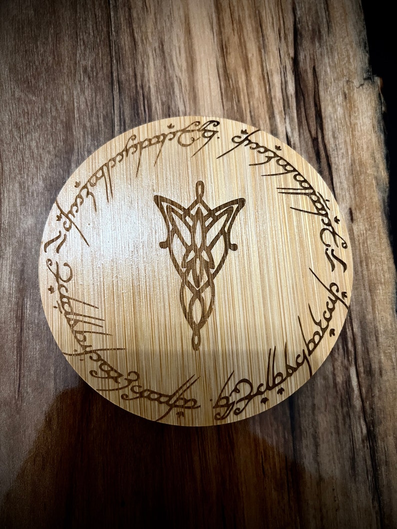Pode incluir: Um descanso de copo redondo de madeira com acabamento em gr&atilde;o de madeira castanho. O descanso de copo apresenta um design gravado a laser do Um Anel de O Senhor dos An&eacute;is, com a inscri&ccedil;&atilde;o "Um Anel para govern&aacute;-los a todos, Um Anel para encontr&aacute;-los, Um Anel para traz&ecirc;-los a todos e nas trevas prend&ecirc;-los."