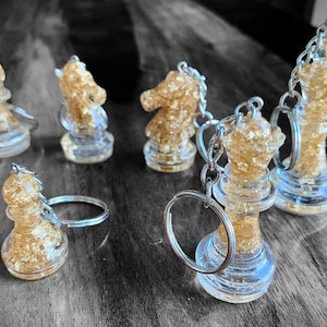 Op de afbeelding: Sleutelhangers met schaakstukken van heldere hars met goudbladaccenten. De set bevat een loper, een paard, een toren, een dame en een koning. Elk stuk is bevestigd aan een zilveren sleutelring.