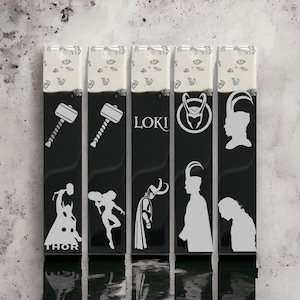 Può includere: Cinque portachiavi rettangolari neri con disegni bianchi. I portachiavi presentano immagini del martello di Thor, dell'elmo di Loki, del viso di Loki e della silhouette di Loki. Sono inclusi anche i testi "LOKI" e "THOR".