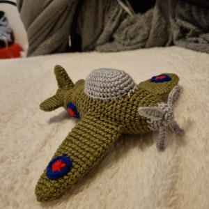 May include: A crocheted green airplane toy with red, white, and blue accents. The airplane has a gray propeller and a gray top.