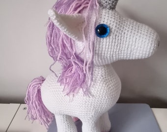 May include: A handmade white crochet unicorn with a gray horn, blue eyes, and lavender mane and tail. The unicorn's hooves are pink. This whimsical stuffed animal is perfect for children or as a decorative item.