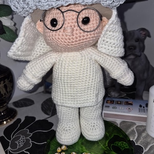 Pode incluir: Boneca de crochê feita à mão usando um traje branco, óculos e um chapéu cinza. A boneca tem um rosto rosa claro e olhos pretos. Está em um prato decorativo verde com enfeites de joaninhas amarelas e pretas.
