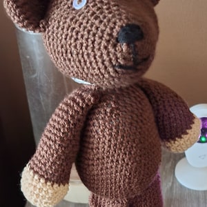 Può includere: Un orsacchiotto di peluche marrone all'uncinetto con occhi a bottone bianchi e un naso nero. L'orsacchiotto è in piedi su una base di legno.