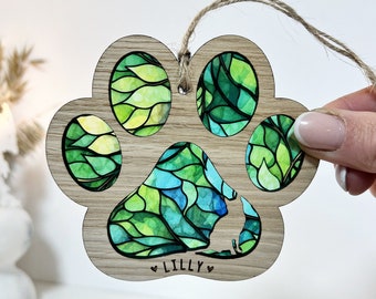 May include: A wooden paw print ornament with a green and blue stained glass design. The ornament has a cutout of a dog in the center and the name "Lilly" is engraved below the cutout.