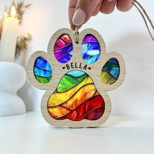 Puede incluir: Un adorno de madera con forma de huella de pata con un diseño de espiral arcoíris y el nombre "BELLA" en el centro.