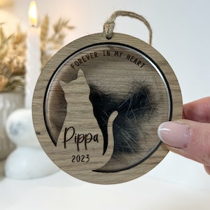 Może przedstawiać: Drewniana ozdoba pamiątkowa dla kota z wygrawerowanymi słowami "FOREVER IN MY HEART" oraz imieniem "Pippa" i rokiem "2023". Ozdoba ma przezroczyste, okrągłe okienko zawierające sierść zwierzęcia.
