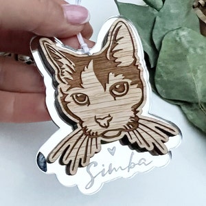 May include: A wooden cat ornament with a mirrored surface. The cat is brown and white with a black nose and whiskers. The ornament is engraved with the name "Simba" and a small heart.