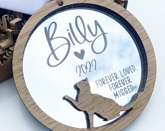 May include: A wooden round ornament with a mirrored surface. The ornament features a cat silhouette, the name "Billy", a heart, and the year "2022". The text "Forever loved. Forever missed" is also engraved on the ornament.