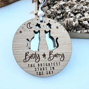 May include: A round wooden ornament featuring two cut-out cats, a crescent moon, and stars. The ornament is personalised with the names "Betty & Barry" and the text "The brightest stars in the sky".