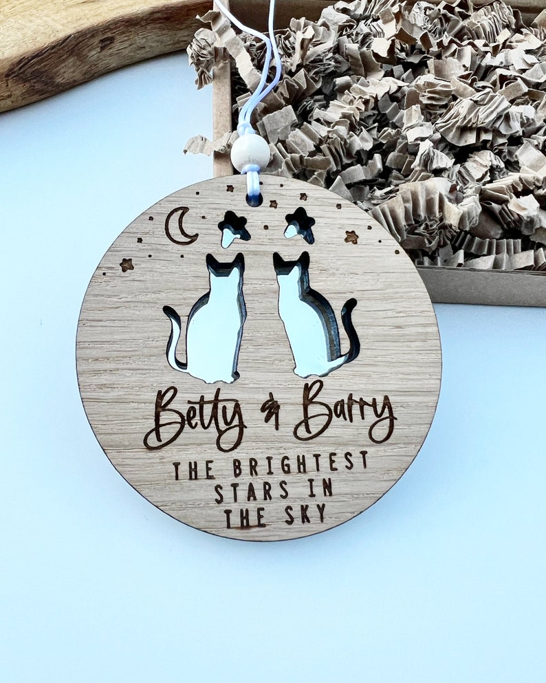 May include: A round wooden ornament featuring two cut-out cats, a crescent moon, and stars. The ornament is personalised with the names "Betty & Barry" and the text "The brightest stars in the sky".