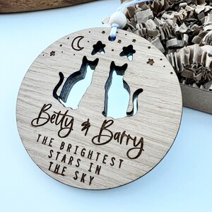 May include: A round wooden ornament featuring two cut-out cats, a crescent moon, and stars. The ornament is engraved with the text "Betty & Barry The Brightest Stars In The Sky".
