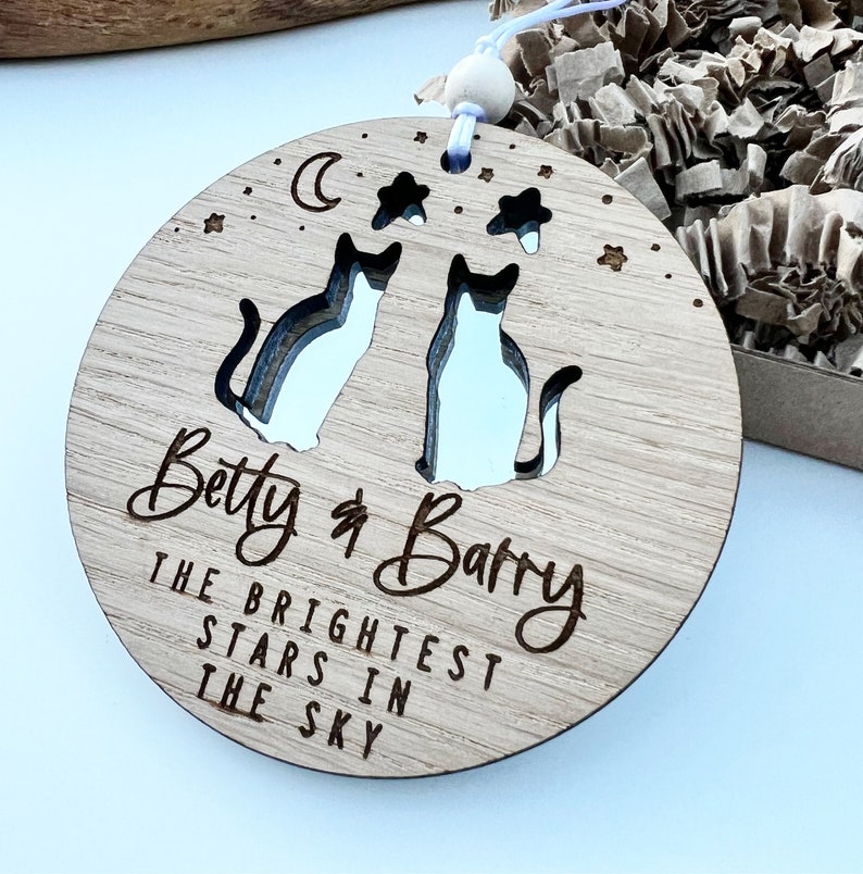 May include: A round wooden ornament featuring two cut-out cats, a crescent moon, and stars. The ornament is engraved with the text "Betty & Barry The Brightest Stars In The Sky".