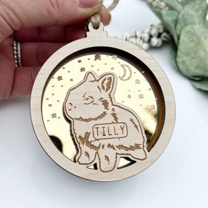 May include: A circular wooden ornament with a gold mirror inside. The ornament features a laser-cut design of a rabbit with the name "TILLY" on a tag. The background of the mirror is gold with white stars.