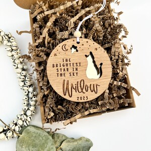 May include: A wooden ornament with a cat silhouette and the text "The Brightest Star in the Sky" and "Willow 2023". The ornament is in a brown box with shredded paper.