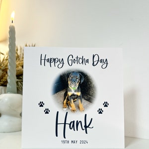 May include: A white card with the text "Happy Gotcha Day" and a photo of a black and brown puppy in a circle. The puppy is sitting on a concrete surface. Below the photo is the name "Hank" in black script and the date "19th May 2024". There are two black paw prints on either side of the name.