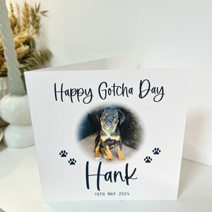 May include: A white card with the text "Happy Gotcha Day" and a photo of a black and brown puppy. The puppy is sitting on a concrete surface. The card also has the text "Hank" and "19th May 2024".
