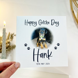 May include: A white card with black text that says "Happy Gotcha Day" and "Hank" with a paw print design. A photo of a black and brown puppy is in the center of the card. The date "19th May 2024" is printed at the bottom of the card.