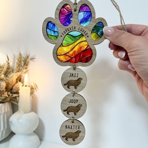 May include: A wooden paw print ornament with a rainbow design and the text "Forever Loved". Three smaller wooden circles hang below with the names "Jazz", "Joop", and "Baxter" and a silhouette of a dog on each.