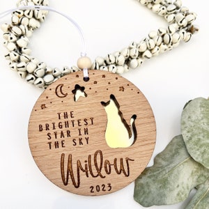May include: A wooden ornament with a cat silhouette and the text "The brightest star in the sky Willow 2023". The ornament is decorated with a crescent moon and stars.