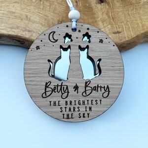 May include: A round wooden ornament featuring a moon, stars, and two cats cut out. The ornament is engraved with the text "Betty & Barry The Brightest Stars In The Sky".