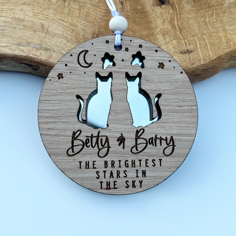 May include: A round wooden ornament featuring a moon, stars, and two cats cut out. The ornament is engraved with the text "Betty & Barry The Brightest Stars In The Sky".
