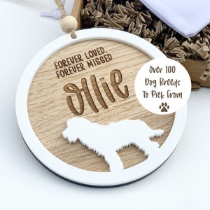 Può includere: Un ornamento rotondo in legno con un bordo bianco, che presenta una silhouette di cane e il testo "Forever Loved, Forever Missed" e un nome personalizzato "Ollie". L'ornamento è accompagnato da un piccolo cartellino bianco con il testo "Over 100 Dog Breeds To Pick From".